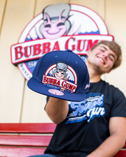 male model holding blue cap with bubba gump logo embroidered. in background is a bubba gump logo on wood wall.
