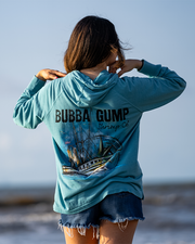 girl standing in front of beach with hands on neck. she is wearing denim shorts and Blue hoodie with back graphic showcasing a boat with compass design behind it. Above graphic reads "Bubba Gump Shrimp Co."