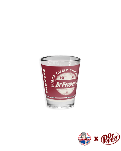 A clear, cylindrical shot glass with a glossy finish, featuring a vibrant red label around its center. The label showcases the iconic Dr Pepper logo in bold white lettering, set against a stylized circular backdrop. Above the logo, ‘BUBBA GUMP SHRIMP CO.’ is prominently displayed, indicating a unique collaboration. Bottom right corner of image includes co-branded logos, suggesting a collectible or promotional item.