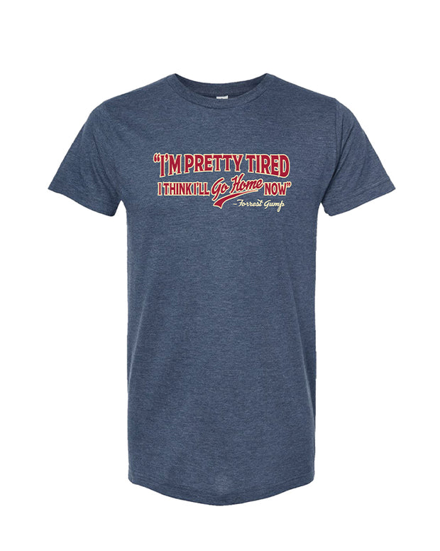 The image shows a heathered blue t-shirt with a graphic text design on the front. The text is in a stylized font, primarily in red with white and blue outlines. It reads: “I’M PRETTY TIRED I THINK I’LL GO HOME NOW -Forrest Gump.”