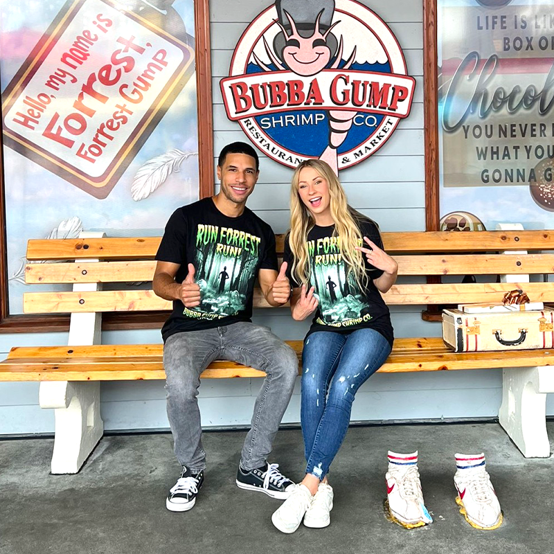  Two individuals are seated on a bench in front of the Bubba Gump Shrimp Co. restaurant. The person on the left is wearing a black t-shirt with green text and the person on the right is wearing a black t-shirt with black text. In front of them is a wooden table with what appears to be a box of chocolates and a sign that reads “Life is like a box of chocolates… you never know what you’re gonna get.” 