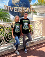 Two individuals are standing in front of a large, rotating globe with the word “UNIVERSAL” on it, indicative of the Universal Studios theme park. Both are wearing matching black t-shirts with the text “RUN FORREST RUN!” and an image that appears to be related to the movie Forrest Gump. They are leaning against a white ornate railing, and behind them is a clear blue sky with some clouds.