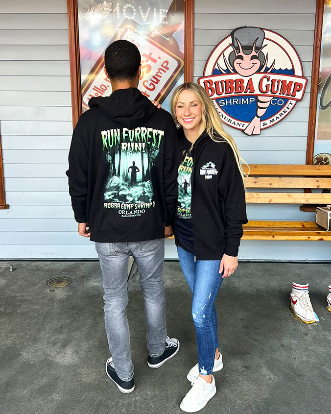 Two individuals are standing in front of a Bubba Gump Shrimp Co. restaurant. The person on the left is wearing a black hoodie with the text “RUN FORREST RUN” and an image of Forrest Gump running, along with the Bubba Gump Shrimp Co. logo and location name “Orlando.” In the background, there’s a wooden bench with a pair of shoes on it, resembling those worn by Forrest Gump in the movie. 