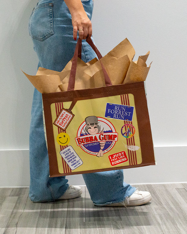 A person is holding a large paper shopping bag with handles. The bag has a colorful design featuring the “Bubba Gump Shrimp Co.” logo prominently displayed in the center, with a red background and a shrimp illustration. Surrounding the logo are various decorative elements, including a yellow smiley face, text bubbles with phrases like “RUN FORREST RUN” and “I JSUT FELT LIKE RUNNING’,” and a feather. The person is wearing light blue denim jeans and white sneakers, standing against a plain grey background.