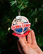 A round ornament with the "BUBBA GUMP SHRIMP CO." logo and a cartoon shrimp character wearing a white top hat. A red banner across the bottom reads "ANAHIEM, CA". The ornament is attached to a Christmas tree and is being held up by a person's hand with green nail polish.