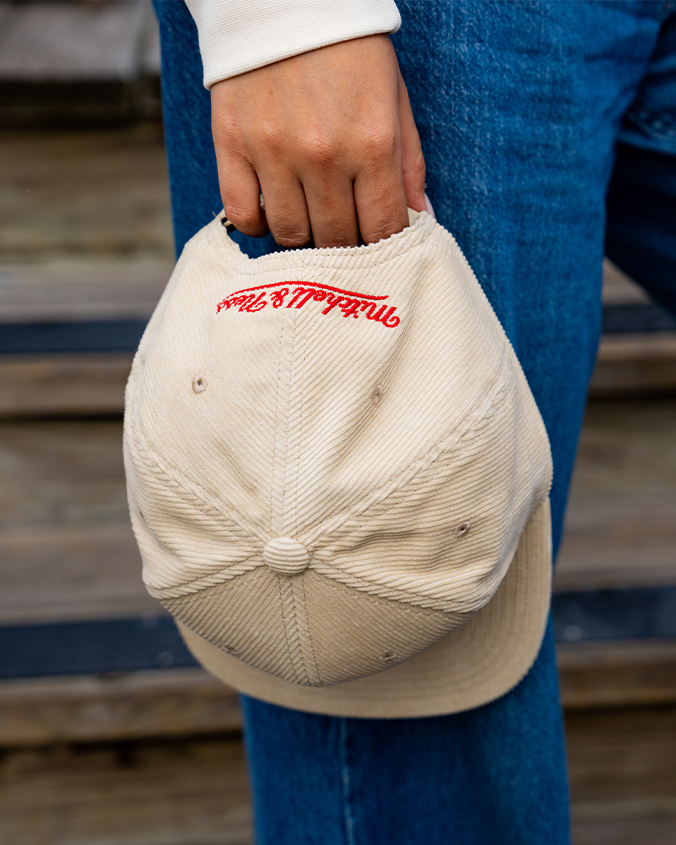 A person holding a beige baseball cap by the back strap. The person is wearing blue jeans and a white long-sleeve shirt. The cap has red embroidered cursive text reading "Mitchell & Ness".