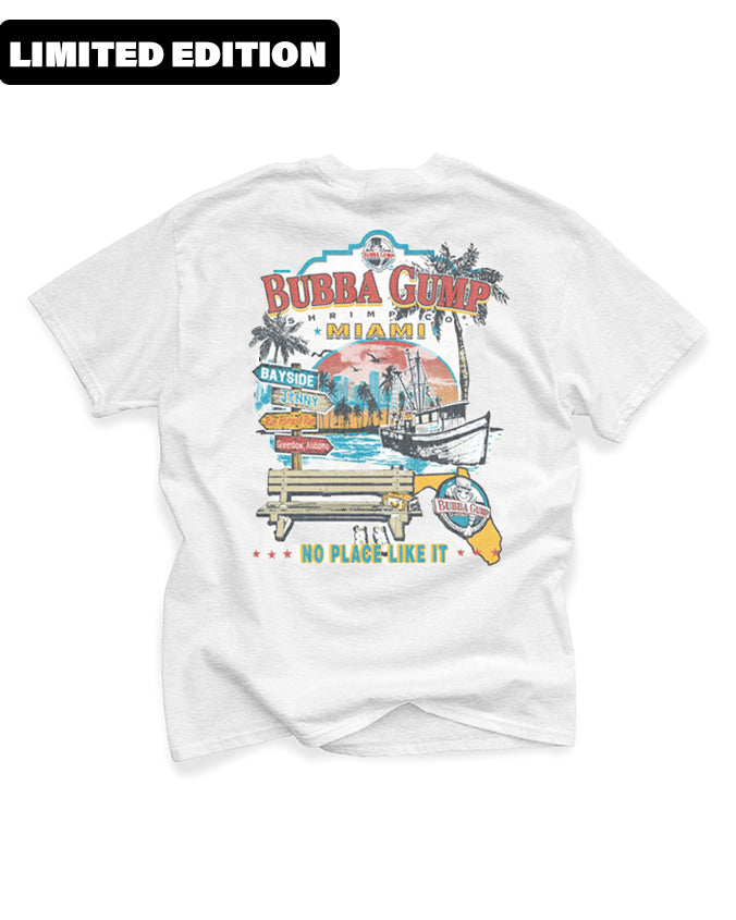 A white t-shirt with a colorful graphic print on the back. The print features the text ‘LIMITED EDITION’ at the top in red, followed by ‘BUBBA GUMP’ in large, bold letters with a shrimp illustration above it. Below is an image of a boat on water with ‘BAY SIDE MIAMI’ written underneath and ‘NO PLACE LIKE IT’ at the bottom of the design. The overall theme suggests a souvenir shirt from Bubba Gump Shrimp Co., highlighting its association with Miami’s bay side area.