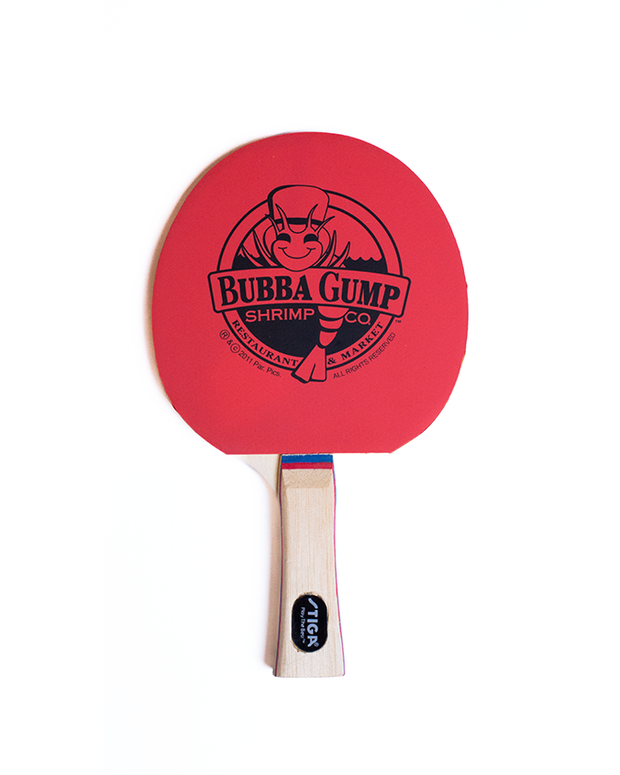 Red ping pong paddle with ‘BUBBA GUMP’ logo in black, wooden handle. On a white background. 