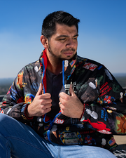 male model sitting down, looking to the side, holding jacket open. puffer jacket is black with drawings of chocolate boxes, ping pong paddles, white feather, muddy smiley face, bubba gump logo, tie dye peace sign.