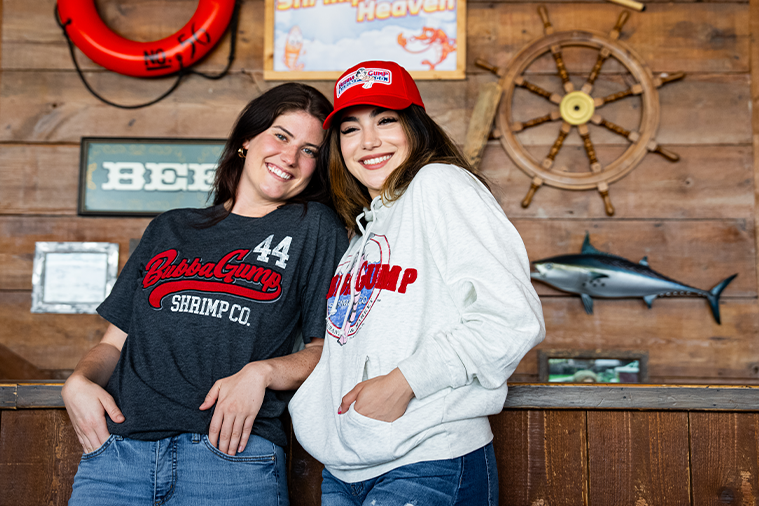 two female models inside a bubba gump restaurant. the background is all wood with nautical wall decor. left model is wearing Charcoal tee with red wording in center that says "Bubba Gump". Right model is wearing red Authentic Bubba Gump Cap & Oatmeal hoodie with bubba gump logo in center.