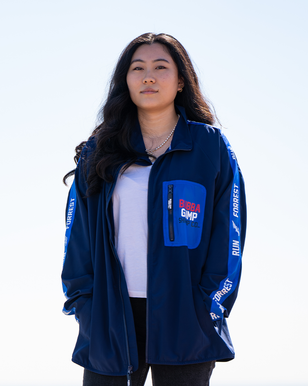 A person wearing a blue zip-up jacket with various text , including 'BUBBA GUMP SHRIMP CO' on the left size of the zip-jacket and 'RUN FUREST RUN' text on the sleeves, paired with a white shirt, against a clear sky background.