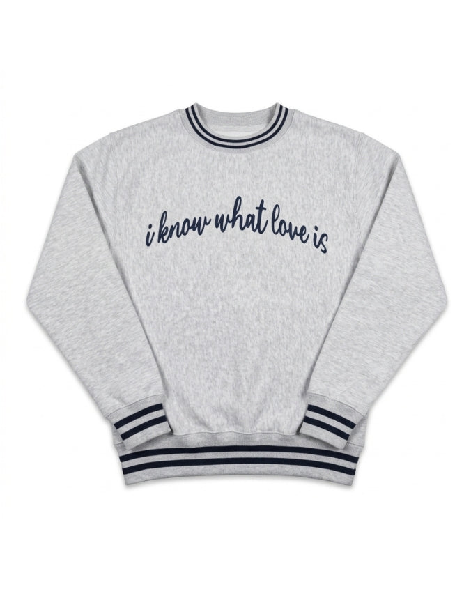 A light heather gray crewneck sweatshirt with navy blue text reading 'i know what love is' across the chest, finished with navy blue striped ribbing on the collar, cuffs, and hem.