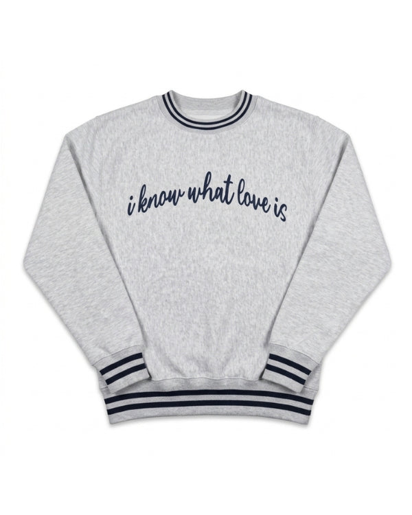 A light heather gray crewneck sweatshirt with navy blue text reading 'i know what love is' across the chest, finished with navy blue striped ribbing on the collar, cuffs, and hem.