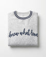 A folded light heather gray sweatshirt with navy blue trim on the crew neck and navy blue script text reading 'I know what love' across the front, shown on a clean white background.