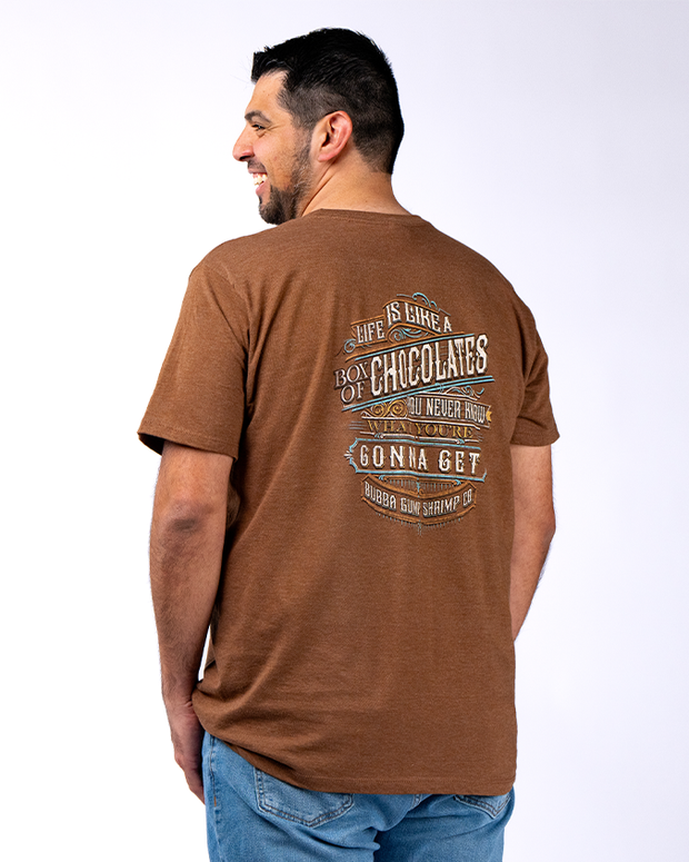 A person wearing a brown t-shirt with a quote from the movie Forrest Gump printed on the back, paired with blue jeans, standing against a white background. The quote on the t-shirt reads: ‘Life is like a box of chocolates you never know what you’re gonna get - Bubba Gump Shrimp Co.’ The text is printed in white colors making it stand out against the brown background of the shirt.