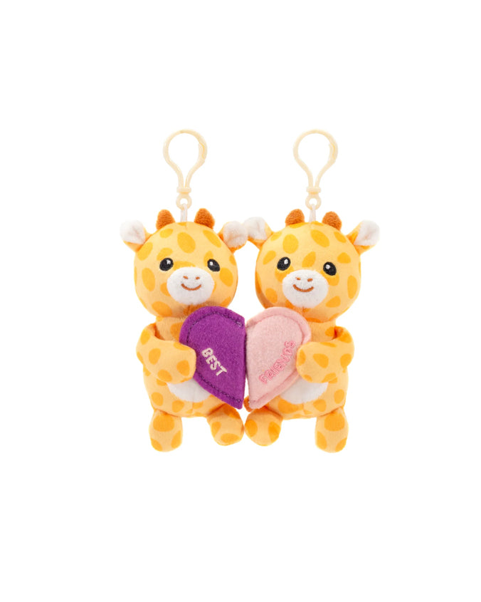 Two plush giraffe toys with smiling faces connected by a magnetic heart. Each giraffe has orange spots on a cream body, small brown horns, and a tuft of brown hair on top of their heads. They have orange ears with white inner ear detailing and are attached to yellow clips at the top. The heart between them is purple on one side and pink on the other, with the word ‘BEST’ embroidered in white on the purple side and ‘FRIENDS’ on the pink side, symbolizing a best friends connection.