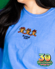 A close-up view of a light blue t-shirt featuring an embroidered design with three colorful frogs and the ‘Rainforest Cafe’ text on the chest. Below, a logo commemorates ‘30 WILD YEARS! Rainforest Cafe’ against a dark green to black gradient background