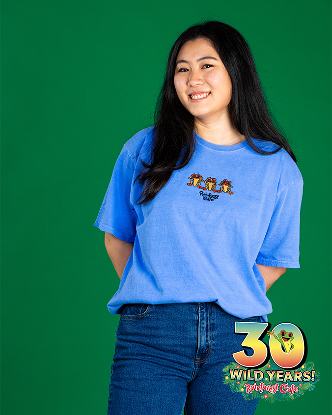 An individual in a blue t-shirt with a graphic of three colorful characters and text on the left side, paired with dark blue jeans, stands against a vibrant green background. The person’s face is not shown. In the bottom right corner, there’s a colorful logo and the text ‘30 WILD YEARS! Rainforest Cafe.