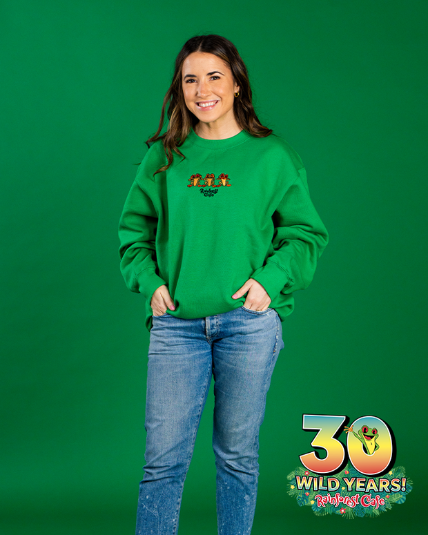 A person stands against a solid green background, wearing a vibrant green crewneck long-sleeve shirt with three frogs embroidered on the center and Rainforest Cafe under them. She isalso wearing blue jeans and have their hands in their pockets. In the bottom right corner of the image, there’s a graphic celebrating ‘30 WILD YEARS! Rainforest Cafe’ with a tree frog coming out the 0.