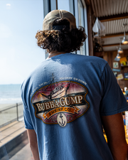 back of male model in restaurant wearing a cap. dark blue short sleeve tee with orange, red, and black graphics on the back. The graphics depict a ship sailing in the sea during sunset. The words "Bubba Gump Shrimp Co." are in the middle of the design. Underneath the words is an anchor within a beige circle. At the top of the graphics, you will find the phrase "We haul'em in, you chow 'em down." 