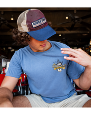 male model sitting down looking down at shirt. Front of the navy overdyes short sleeve scoop neck tee with left chest tiny orange graphics. Graphics depict a small sailing ship and an anchor image at the bottom. In the middle of the graphics, there are the words "Bubba Gump Shrimp Co.