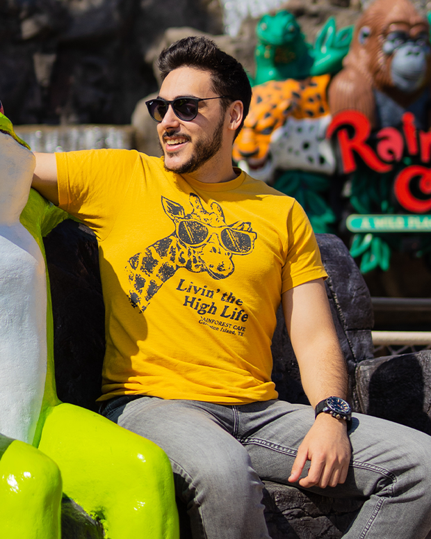 Man in sitting infront of a Rainforest Cafe wearing sunglasses and the "Livin' The High Life" tee. Golden yellow with a close up of a giraffe wearing sunglasses. Reads "Livin' the High Life" and underneath that, Rainforest Cafe.