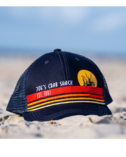 cap on sand. Navy blue cap with Joe's Crab Shack branding and small crab boat being illuminated by moon.
