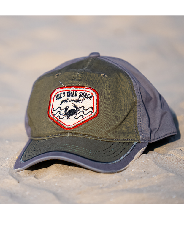 cap on sand. Grey and green two tone cap with patch saying "got crabs?" with crab embroidery and Joe's Crab Shack branding.
