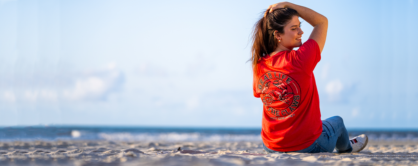 girl sitting on sand. she is holding her hair up with her right arm and facing to the right. She's wearing a red tee shirt with blue jeans and white tennis shoes.