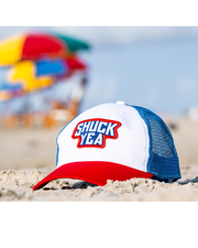 Mesh cap that has the phrase "Shuck Yea' embroidered on the front. back mesh is a navy blue, front is white and the bill is red placed on top of sand with colorful beach umbrellas in background.