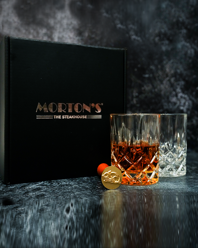 A black box on a black marble table with the text 'MORTON'S THE STEAKHOUSE' on it. In front of the box gold ice stamp with an intricate design and a wooden handle, placed in front of two crystal whiskey glasses, one empty one full.