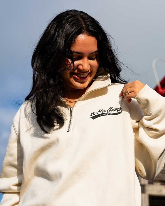 A person wearing a cream-colored zip-up sweatshirt featuring the "Bubba Gump Shrimp Co." logo, with long black hair styled loosely.