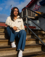 A person sits on wooden steps wearing a cream-colored zip-up sweater, blue jeans, and white sneakers, with a restaurant in the background.