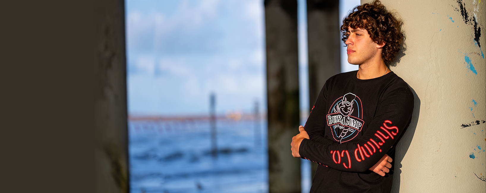 male model with curly hair, wearing black long sleeve Bubba Gump x Mitchell & Ness top. He has his arms crossed and is leaning against a pole at the beach.