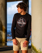 male model leaning against a pillar at the beach. he is wearing Black long sleeve scoop neck tee with neon graphics on the front chest and both sleeves sides. The white, blue, and neon red front graphic is a Bubba Gump Classic logo in neon style. 