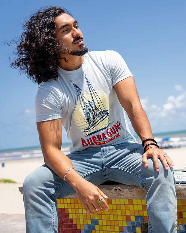 male model with curly hair sitting on bench in front of the beach. He is wearing Light blue crew neck short sleeve tee, fitted on the mannequin. It has boat graphics with navy blue shrimp boat outlines and the large white sun setting on the background of the graphics. At the bottom of the shrimp boat is wording in red, "Bubba Gump" and below that in the red ribbon, "shrimp co."