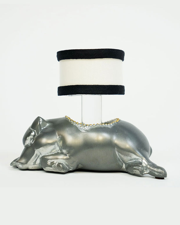 A metallic silver sculpture of a laying pig with a flat surface on its back supporting a cylindrical white lampshade with black bands at the top and bottom. The lampshade is positioned where the pig's back would naturally rise, turning the sculpture into a functional lamp.