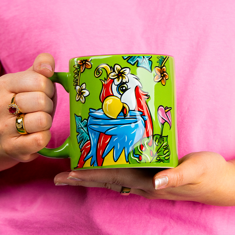 A vibrant green mug adorned with a colorful illustration of a parrot surrounded by tropical flowers, held against a pink shirt. 