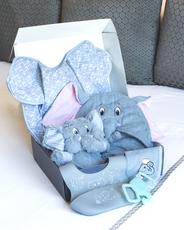 A baby gift box with a blue animal-patterned exterior, containing a soft grey plush elephant toy with pink inner ears, a smaller matching elephant toy, a blue and white zigzag patterned blanket, and a greenish-blue baby teether shaped like keys, all arranged neatly inside the box  on top of a bed with grey pillows and white blanket.