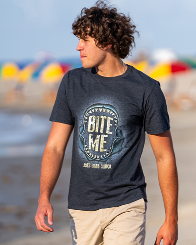 A person stands on a sunny beach, looking to the left, wearing a dark t-shirt with a shark jaw design and the playful text ‘BITE ME’ above it, with colorful beach umbrellas dotting the background.
