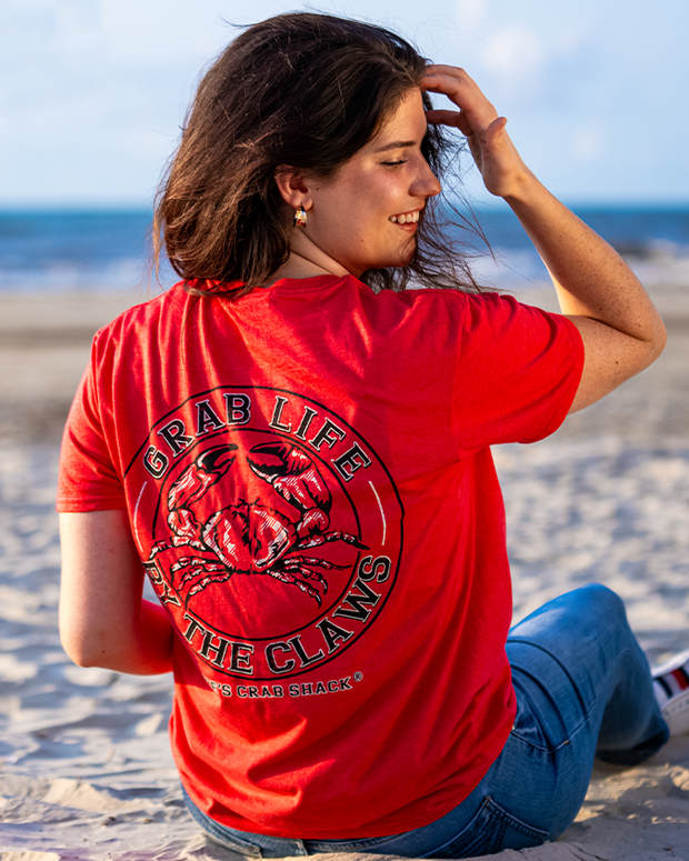 female model sitting on sand at beach. she is looking over her shoulder, smiling. the shirt back graphic is a circle with a crab in the center. words around it read "grab life by the claws".