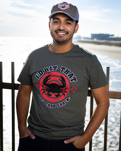 Man standing in front of the beach wearing the "i'd Hit That Tee". Tee is a military green that reads in black letters " I'd Hit That" & "Joe's Crab Shack". In between both lines, an image of a black crab silhouette on red circles.