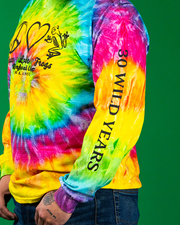 A person stands against a solid green background, wearing a vibrant, multi-colored tie-dye sweatshirt with ‘30 WILD YEARS’ printed on the sleeve. The sweatshirt features a kaleidoscope of colors including yellow, blue, red, and green. The individual has their hands casually placed in the pockets of blue denim jeans.