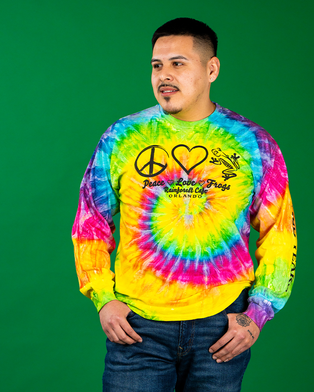 A person stands against a solid green background, wearing a vibrant tie-dye sweatshirt adorned with symbols of peace and love, and the text ‘Peace Love Rainforest Cafe Frogs.’ The colorful patterns swirl in yellow, green, blue, red, and purple, while the person casually places their hands in the pockets of their dark jeans.