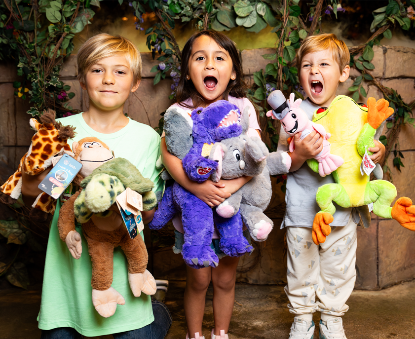 Three children hold colorful plush toys, including a giraffe, monkey, dinosaur, elephant, and frog, surrounded by greenery.