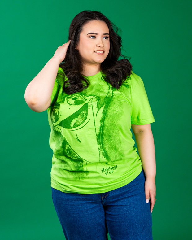 female model is standing in front of green background wearing cha cha shirt and jeans. she has one hand along her ear, fixing her hair and looking off to the side.