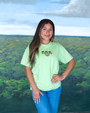 A person stands in front of a painted landscape background, featuring lush green treetops. They are wearing a light green t-shirt with a graphic of three tree frogs and the word ‘rainforest cafe’ written below them in black font. The individual is also wearing blue denim jeans and has one hand resting on their hip. 
