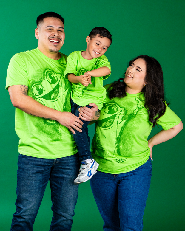 male, female and boy model wearing cha cha tee and denim jeans. all are standing in front of a green background, laughing. male model is holding boy model, while female model is looking at boy.