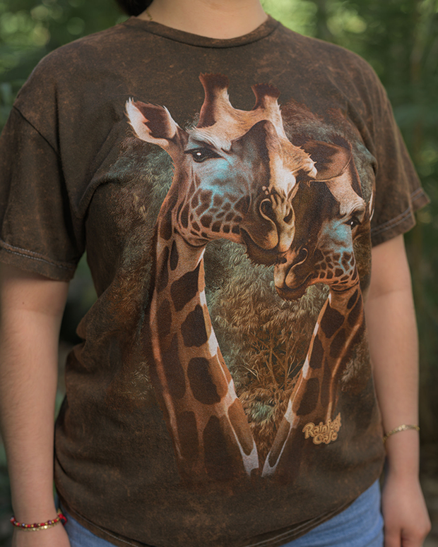 A person wearing a brown t-shirt with a striking graphic of a giraffe’s head and neck, which extends from the bottom hem up towards the wearer’s right shoulder. The giraffe is set against a backdrop of trees and foliage, suggesting a natural habitat. The word “Rainforest Cafe” is printed at the bottom right corner of the shirt.