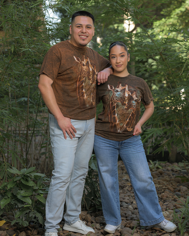 Two individuals standing outdoors on a rocky terrain with greenery in the background. They are both wearing t-shirts adorned with a graphic of a giraffes. The person on the left pairs their shirt with white pants and casually places their left hand in their pocket, while the person on the right complements their shirt with blue jeans.
